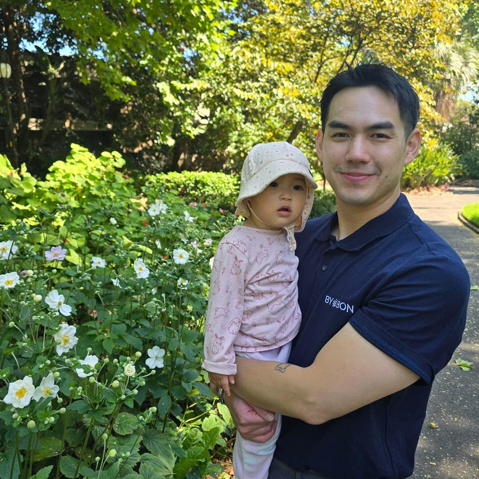 Man holding a child in a park with greenery and flowers in the background